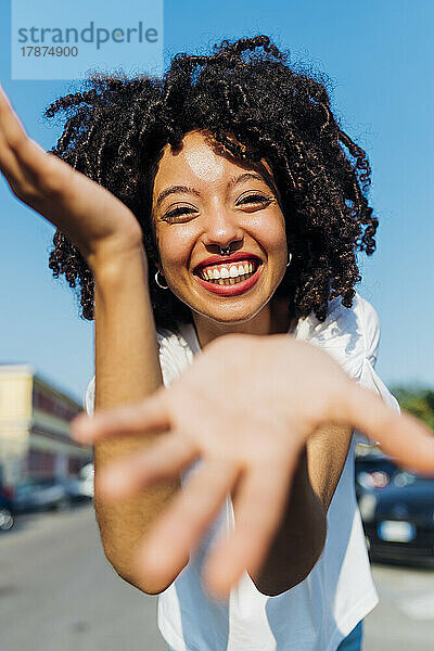Glückliche junge Frau mit lockigem Haar gestikuliert an einem sonnigen Tag