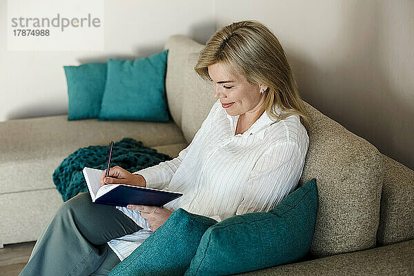 Lächelnde reife Frau schreibt Tagebuch und sitzt zu Hause auf der Couch