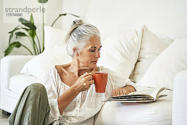 Reife Frau mit Tasse liest Buch im Wohnzimmer