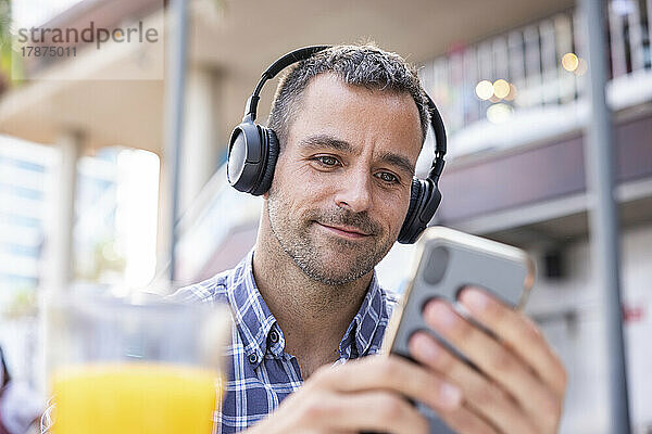 Lächelnder reifer Mann  der im Café sein Smartphone benutzt und über Kopfhörer Musik hört