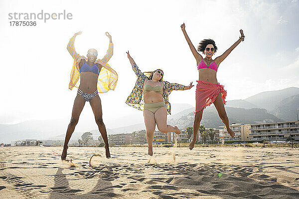 Sorglose Freunde springen an einem sonnigen Tag mit erhobenen Armen am Strand