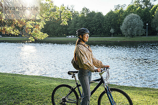 Lächelnde Frau mit Fahrrad steht am See im Park