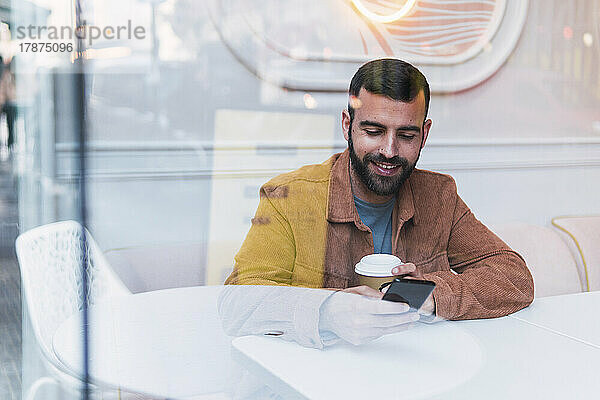 Lächelnder junger Mann benutzt Smartphone  gesehen durch Glas im Café