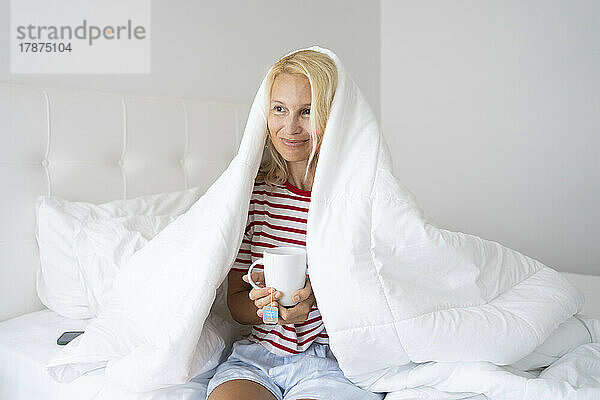Lächelnde Frau hält in eine Decke gewickelten Tee auf dem Bett
