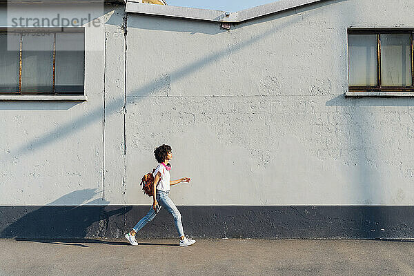 Junge Frau mit Rucksack geht auf Fußweg an Mauer entlang