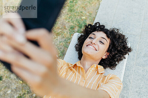 Glückliche junge Frau macht ein Selfie mit ihrem Smartphone  das auf einer Bank liegt