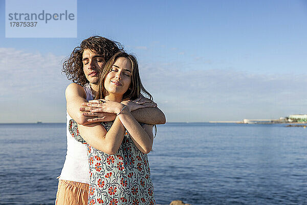 Junges Paar steht an einem sonnigen Tag mit den Armen vor dem Meer