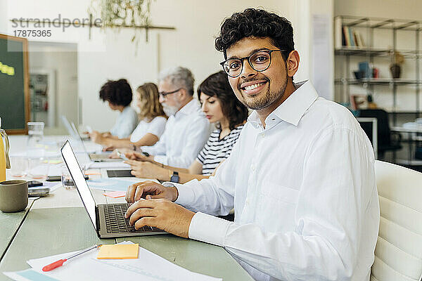 Lächelnder Geschäftsmann mit Laptop sitzt neben Kollegen im Büro