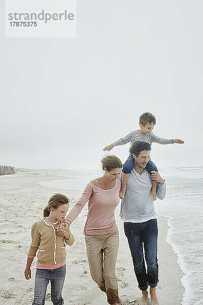 Eltern gehen mit zwei Kindern am Strand spazieren und tragen ihren Sohn auf den Schultern