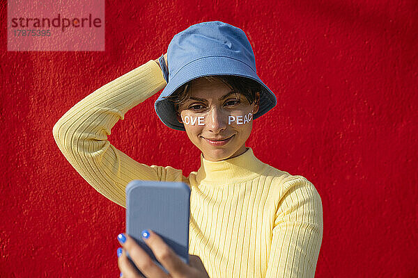 Frau mit Fischerhut macht Selfie mit Smartphone vor roter Wand