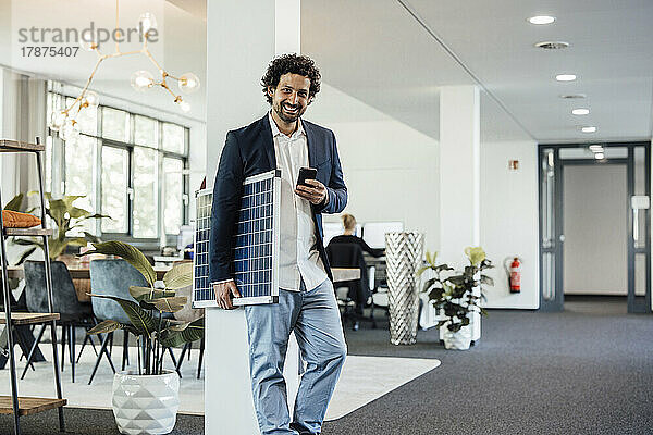 Lächelnder Geschäftsmann mit Smartphone und Solarpanel im Büro