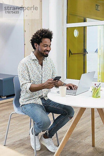 Lächelnder Geschäftsmann mit Mobiltelefon und Laptop im Büro
