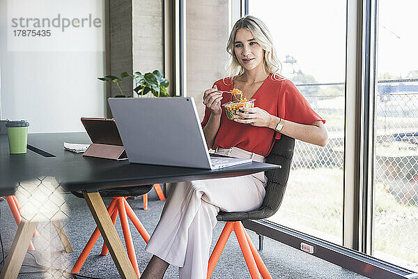 Geschäftsfrau isst Salat und schaut auf den Laptop  während sie im Büro auf einem Stuhl sitzt
