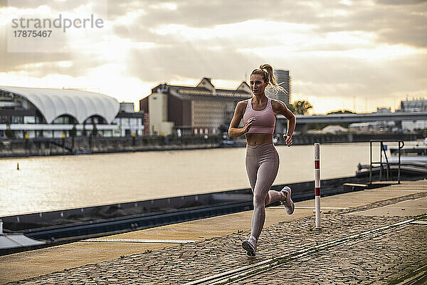 Blonde Frau joggt bei Sonnenuntergang vor dem Fluss