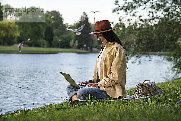 Lächelnde Frau mit Laptop sitzt am See im Park