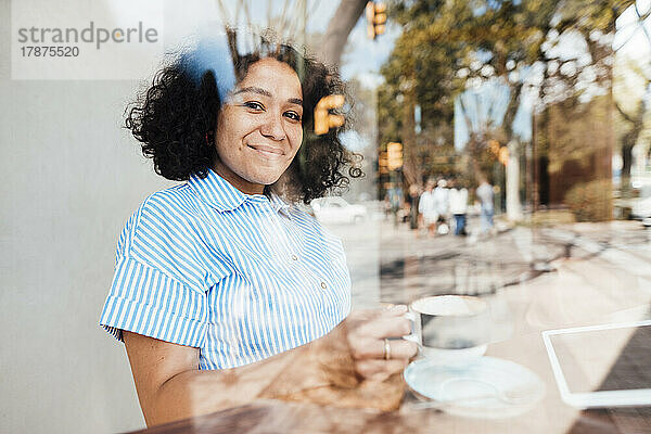 Lächelnde Frau mit Kaffeetasse  gesehen durch ein Glas Café