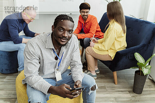 Lächelnder Geschäftsmann hält Smartphone mit Kollegen im Hintergrund im Büro