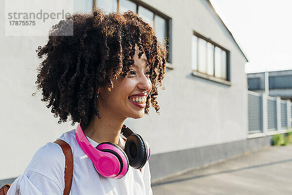 Glückliche junge Frau mit lockigem Haar  die an einem sonnigen Tag kabellose Kopfhörer trägt