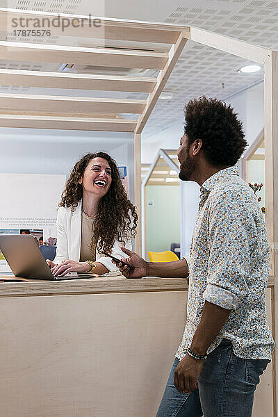 Lachende Geschäftsfrau mit Laptop blickt Kollegen im Büro an