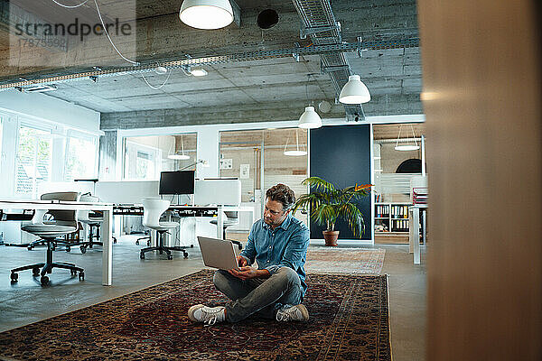 Businessman using laptop sitting on carpet in office