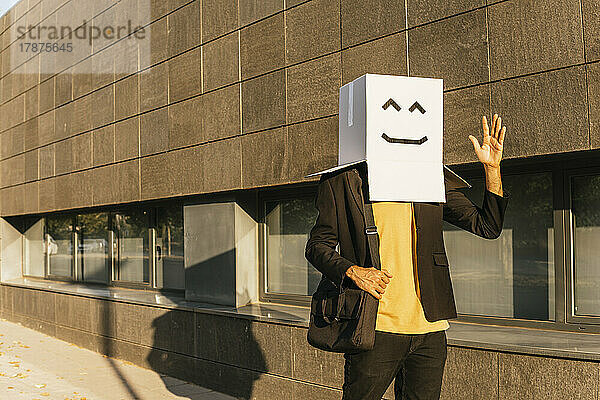 Geschäftsmann trägt Box mit Smiley-Gesicht und winkt an einem sonnigen Tag auf dem Fußweg