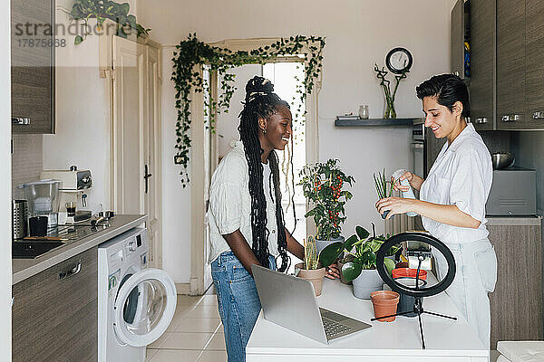 Lächelnde junge Frau im Gespräch mit einer Freundin  die zu Hause Pflanzen gießt