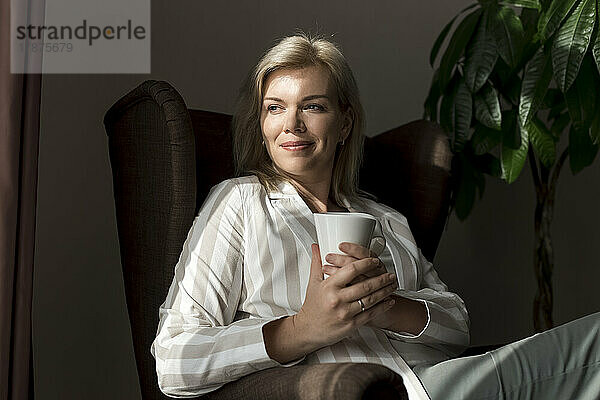 Lächelnde reife Frau mit Kaffeetasse sitzt zu Hause im Sessel