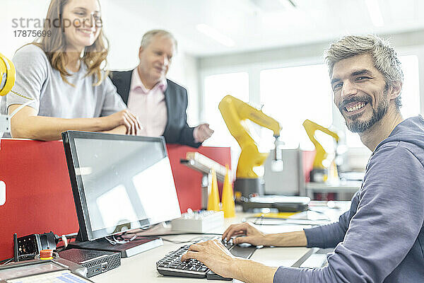Zufriedene Kollegen beobachten lächelnden Mann bei der Arbeit am PC in der Roboterfabrik