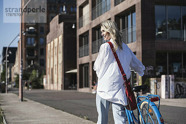 Lächelnde Frau  die an einem sonnigen Tag mit dem Fahrrad vor dem Gebäude läuft
