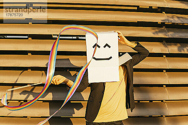 Mann in Smiley-Box dreht an einem sonnigen Tag ein regenbogenfarbenes Band für rhythmische Gymnastik