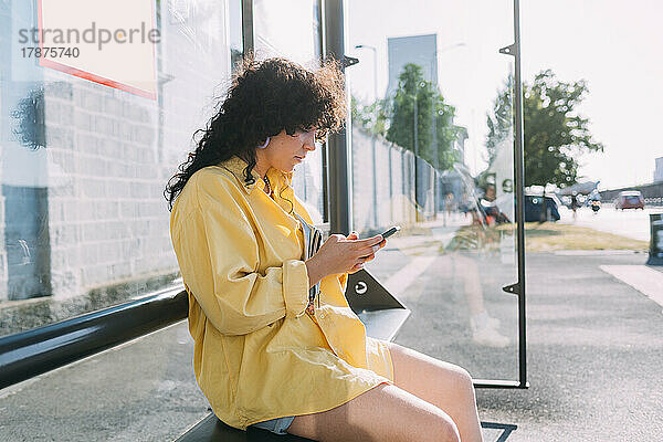 Junge Frau sitzt an Bushaltestelle und benutzt Smartphone