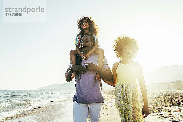 Glücklicher Mann  der ein Mädchen auf der Schulter trägt und an einem sonnigen Tag mit seiner Tochter am Strand spaziert