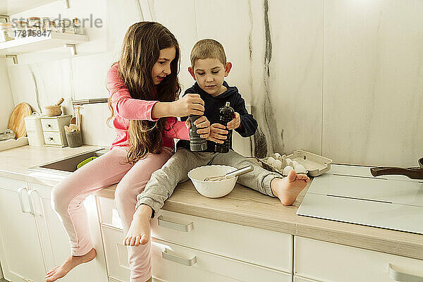 Mädchen und Bruder halten Shaker über Schüssel und sitzen zu Hause auf der Küchentheke