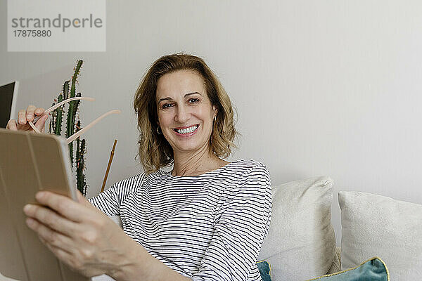 Lächelnde Frau mit Tablet-PC sitzt zu Hause auf der Couch