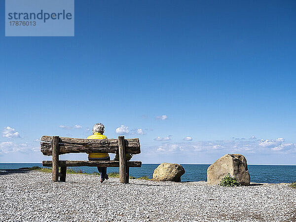 Ältere Frau sitzt auf einer Bank an Felsen am Meer