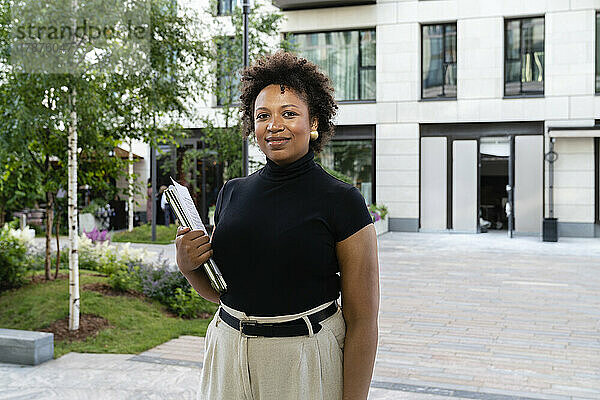 Lächelnde Geschäftsfrau mit Laptop steht vor Bürogebäude