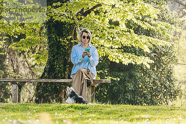 Frau mit Sonnenbrille und Smartphone sitzt auf Bank neben Hund im Park