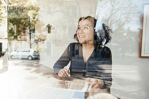 Lächelnde Frau mit Tablet-PC  gesehen durch das Glas eines Cafés