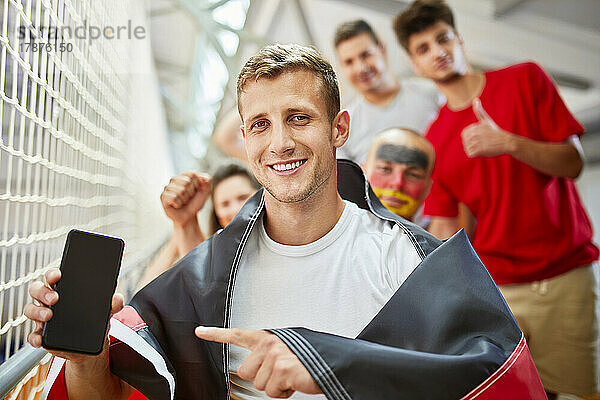 Lächelnder Mann mit deutscher Flagge zeigt auf Smartphone-Bildschirm bei Sportveranstaltung im Stadion