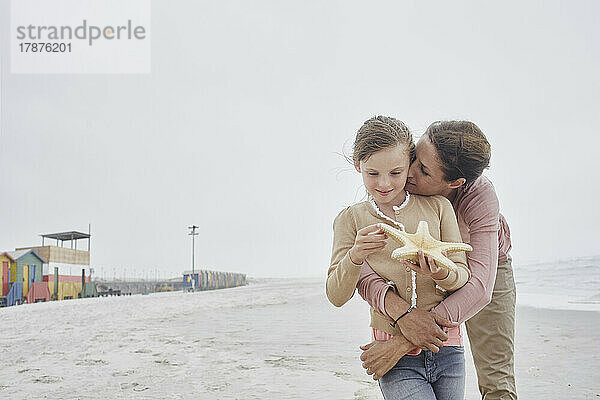 Mutter küsst Tochter am Strand und hält Seesterne in der Hand