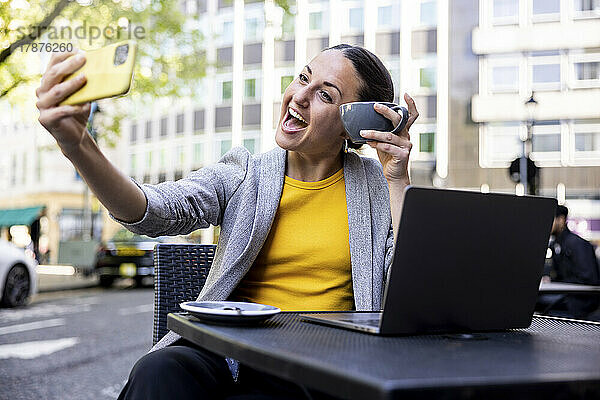 Glückliche Geschäftsfrau hält eine Kaffeetasse in der Hand und macht ein Selfie per Smartphone im Straßencafé