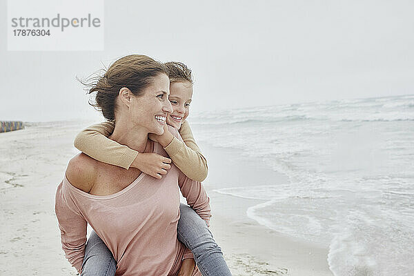 Mutter spaziert am Strand und trägt ihre Tochter huckepack