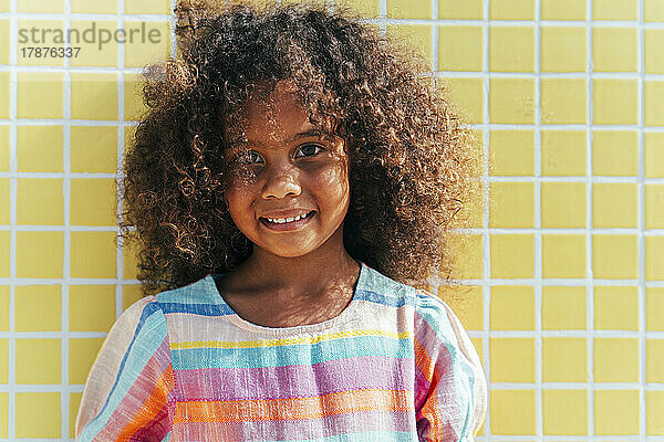 Lächelndes Mädchen mit lockigem Haar vor gelber Wand