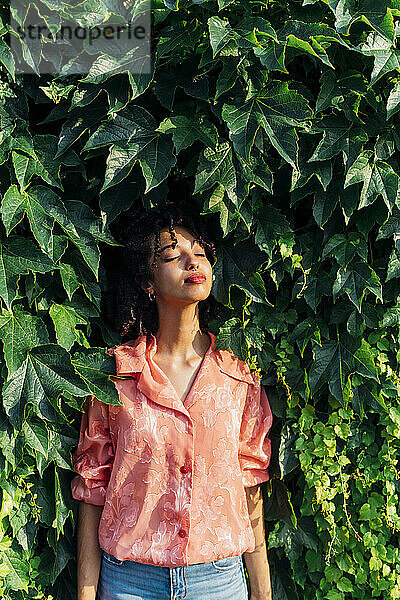 Woman with eyes closed standing amidst green leaves enjoying sunlight