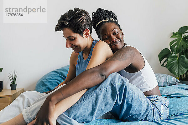 Smiling woman embracing girlfriend from behind sitting in bedroom