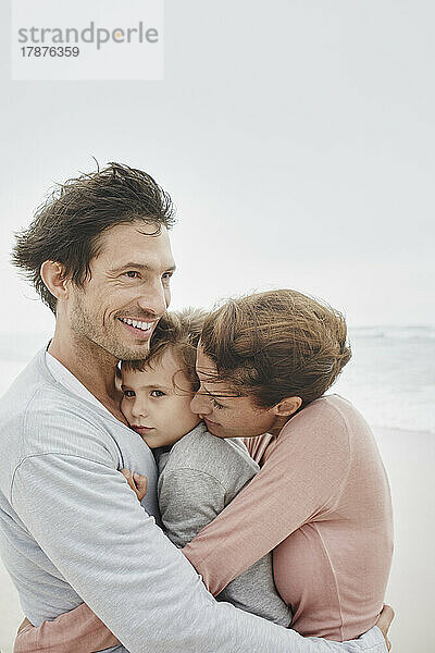 Fürsorgliche Eltern umarmen und küssen ihren Sohn am windigen Strand