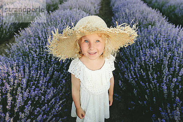 Lächelndes süßes Mädchen mit Strohhut  das im Lavendelfeld steht