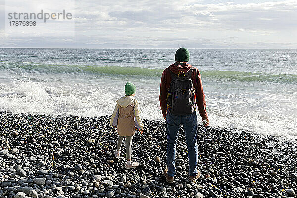 Vater und Tochter stehen am Kiesstrand und blicken auf das Meer