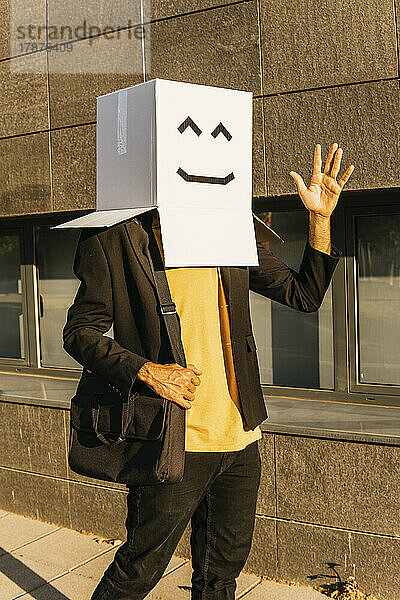 Businessman wearing box with smiley face waving hand on footpath