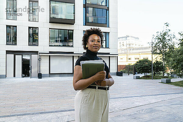 Lächelnde junge Geschäftsfrau mit Laptop vor Bürogebäude
